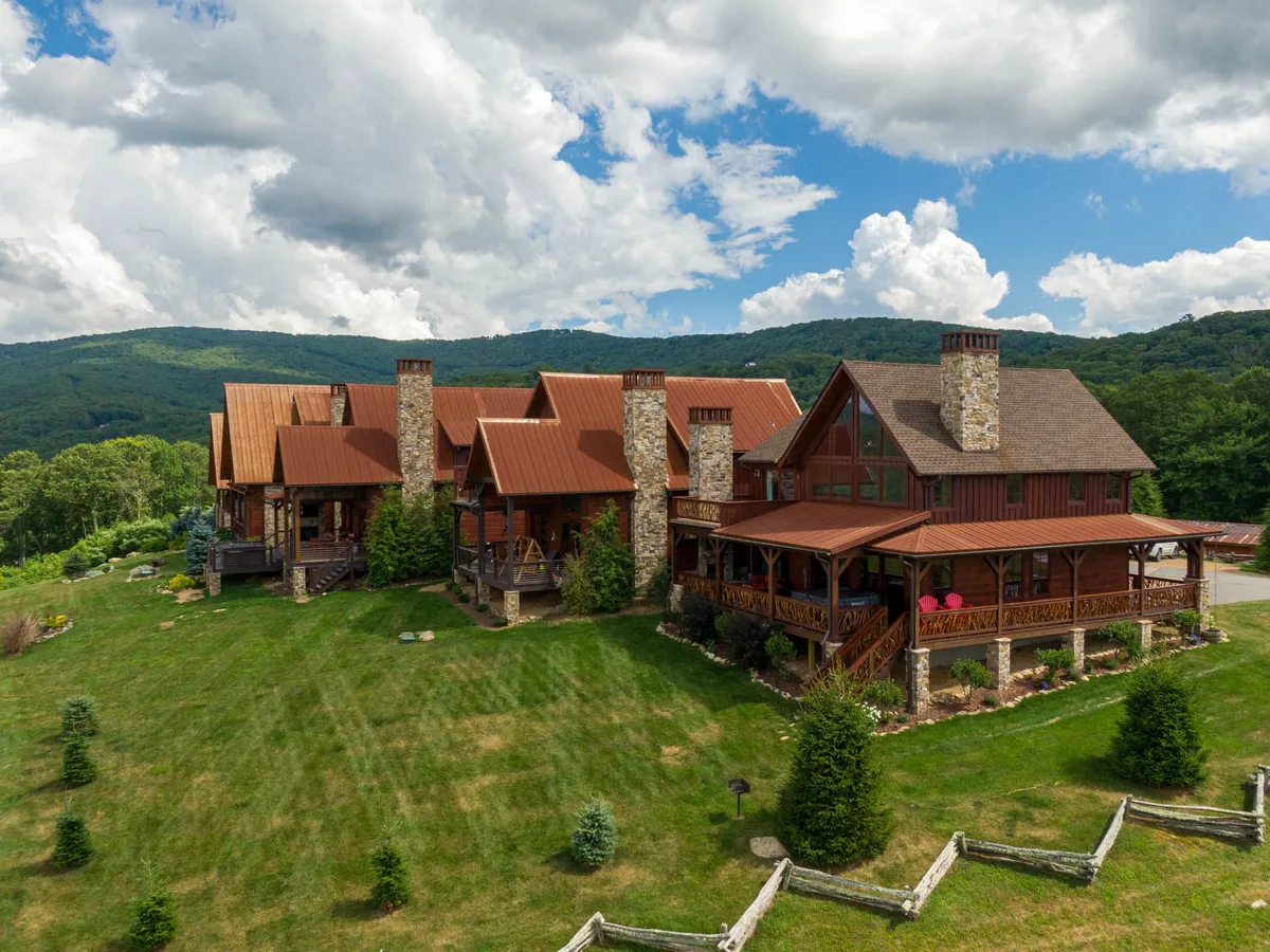 A welcoming exterior shot of the main lodge structure, featuring charming stone chimneys, a spacious wooden deck, and beautifully landscaped gardens. Lush green surroundings and mountain views set a tranquil scene, while the deck suggests a perfect place for gatherings or relaxation.