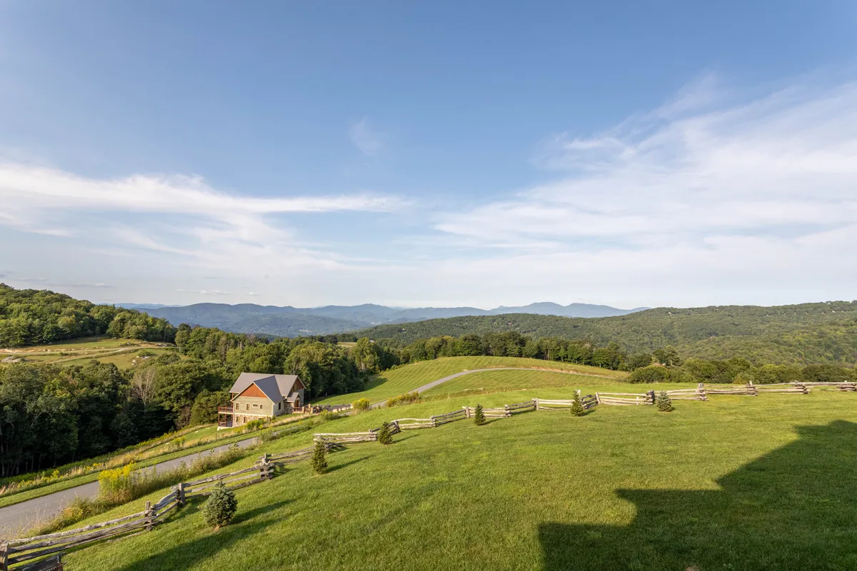 A tranquil scene featuring rolling green hills, distant blue-tinged mountains, a rustic wooden fence, and a house nestled in nature. The open space suggests a perfect setting for outdoor activities like hiking, picnicking, and stargazing.