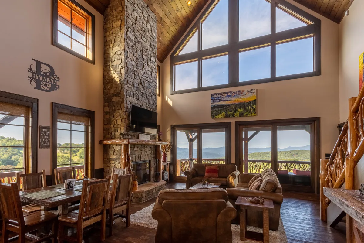 This image showcases a spacious living room with a tall stone fireplace, large windows that frame sweeping mountain views, a wooden dining table, comfortable seating, and tasteful artwork. The natural light and open layout create a welcoming environment perfect for gatherings.