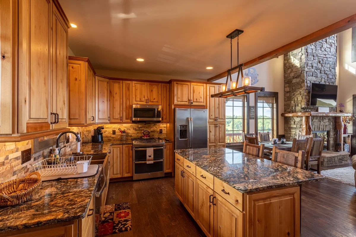 A modern kitchen with a large granite island, warm wood cabinetry, and a view into a cozy living space anchored by a stone fireplace. The setup is perfect for preparing meals together or enjoying casual gatherings at the island bar.