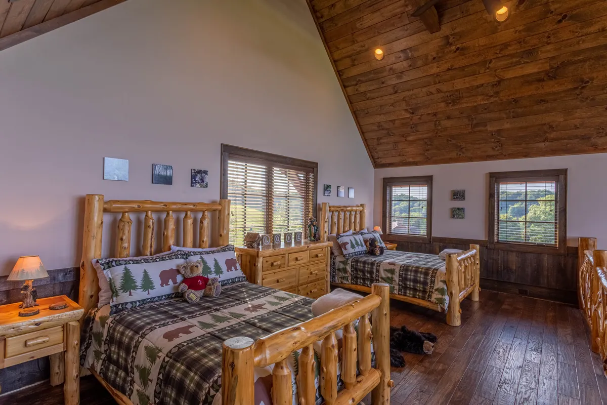 A cozy loft area with rustic log beds, plaid bedding, wooden furniture, and windows with blinds. The space is inviting and flexible, ideal for kids or teens to relax or gather.