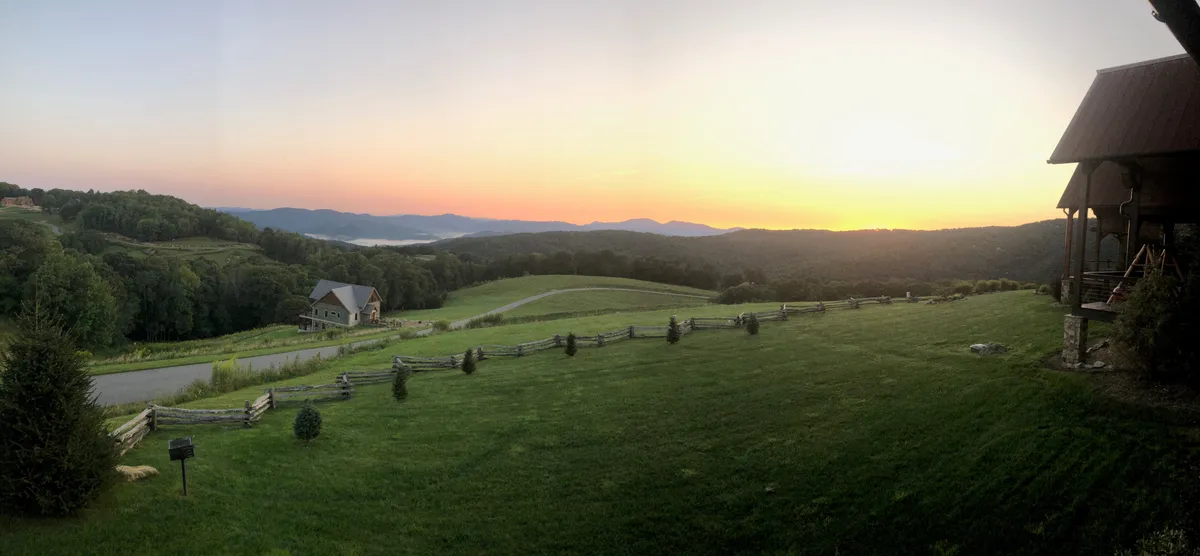 A panoramic view over a mountain range at sunset, with a green lawn and part of a house and fence in the foreground. The scene conveys an expansive sense of privacy, natural beauty, and the opportunity to enjoy stunning sunsets from the property.