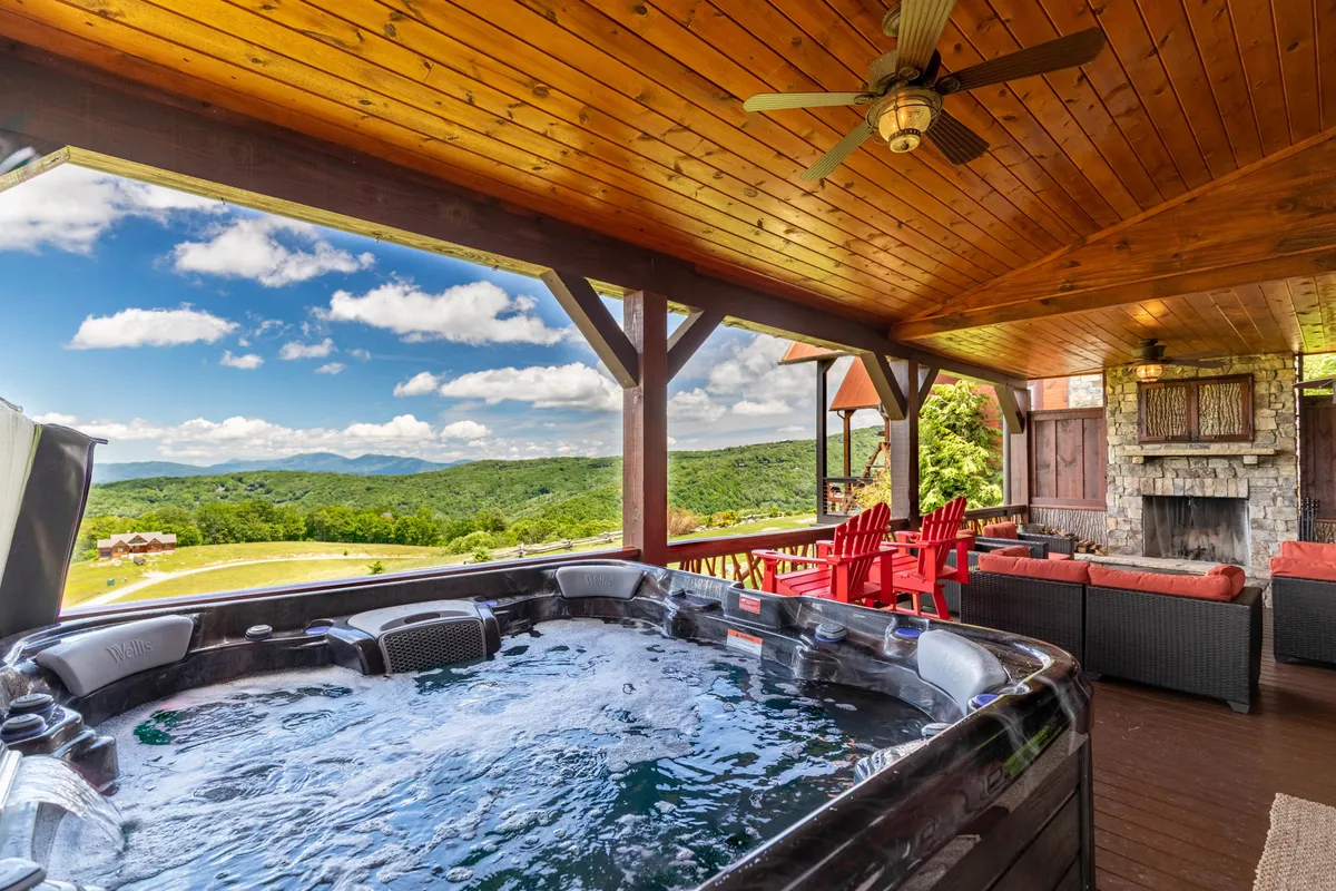 This exterior image features a luxurious hot tub situated on a covered deck, complete with an inviting seating area and a fireplace. The scene is set against a dramatic mountain view, creating an atmosphere of relaxation and escape. The overall setup embodies a perfect blend of comfort and nature, ideal for unwinding at any time of day.