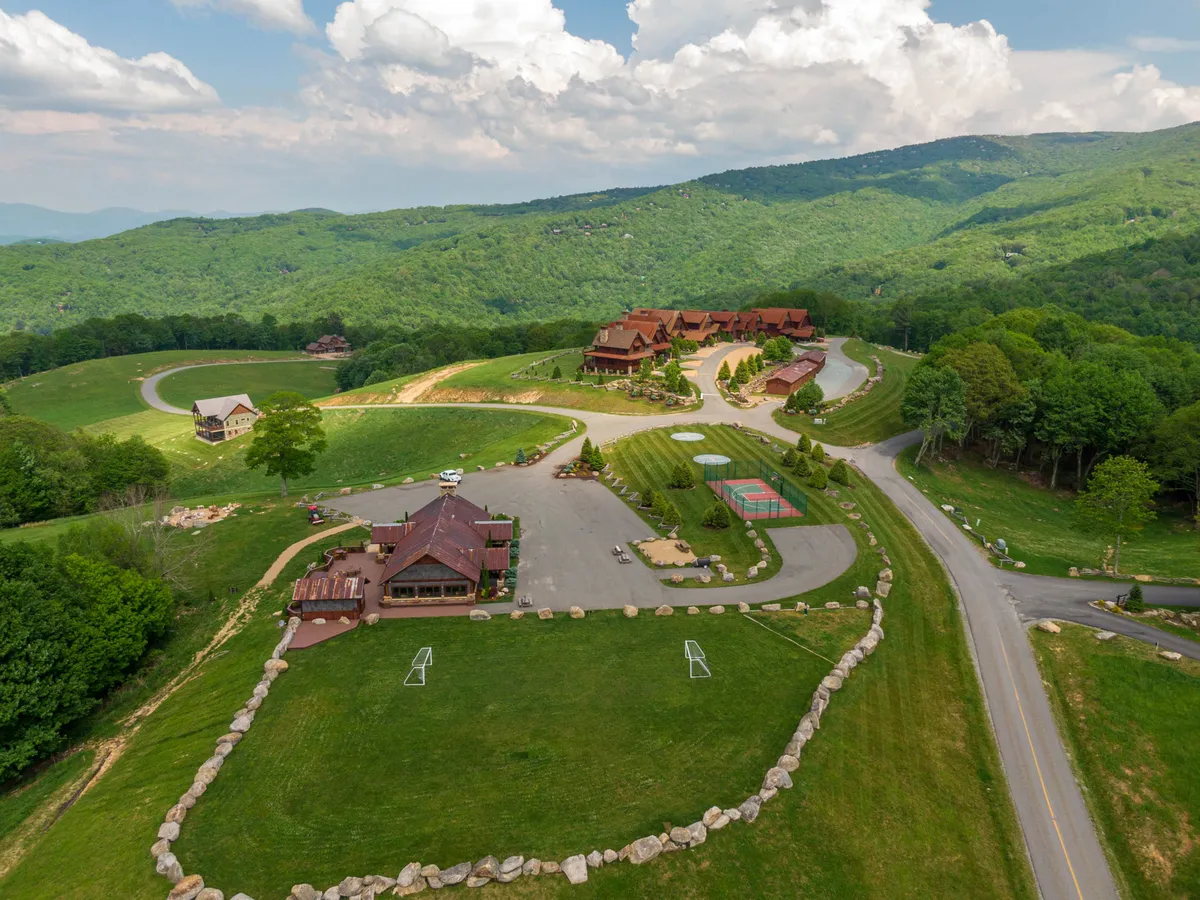 This exterior image shows the main lodge set against rolling hills with a visible sports area and a parking lot in the foreground. The landscape is open and welcoming, with clear pathways leading toward the activity areas, hinting at the variety of outdoor amenities available to guests. The setting evokes a sense of community and the possibilities for sports, gatherings, and exploration right at your doorstep.