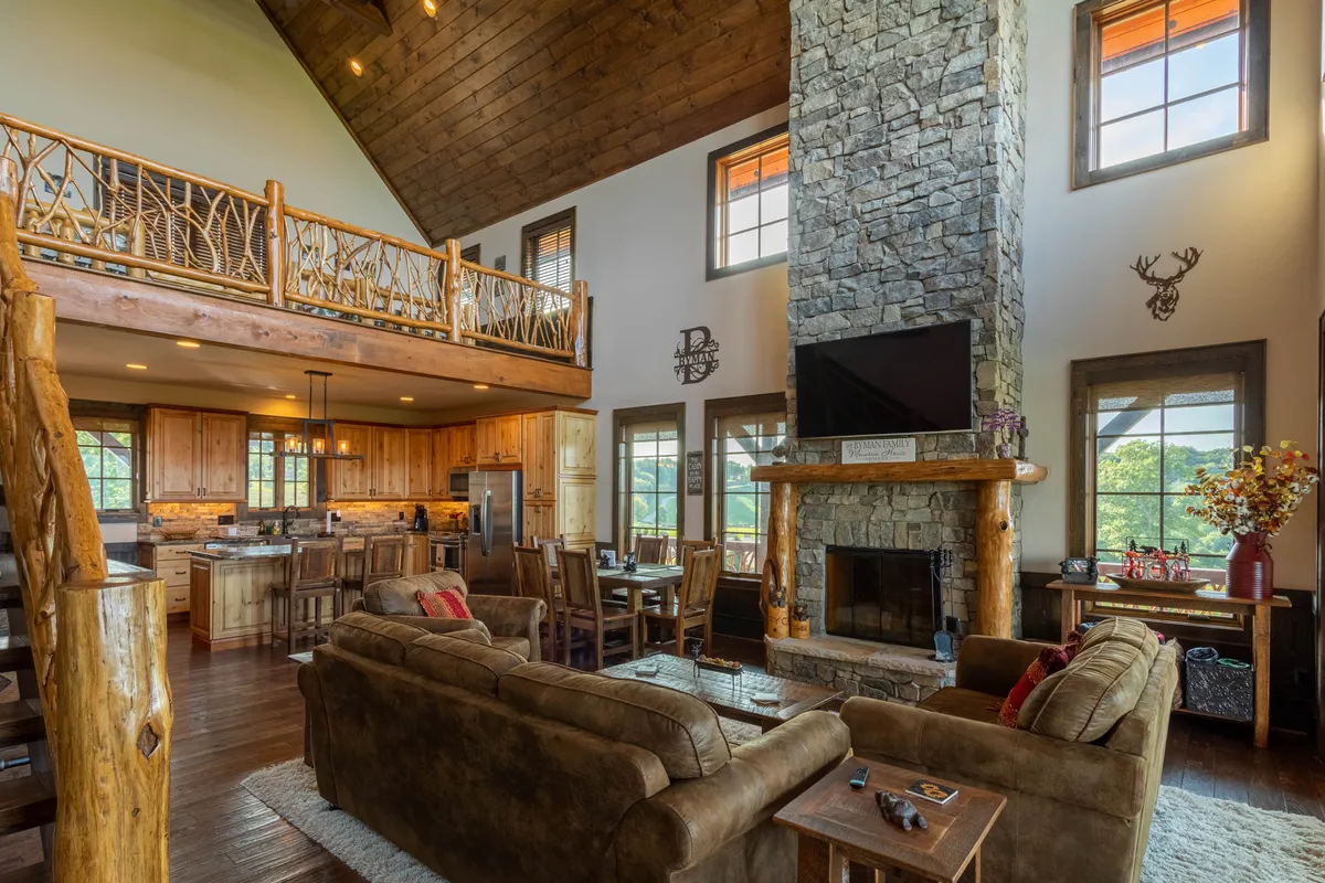A wide view of an inviting great room featuring a stone fireplace, exposed wooden beams, an open kitchen with bar seating, and floor-to-ceiling windows looking out onto natural scenery. The seamless flow between kitchen, living, and dining spaces is apparent.