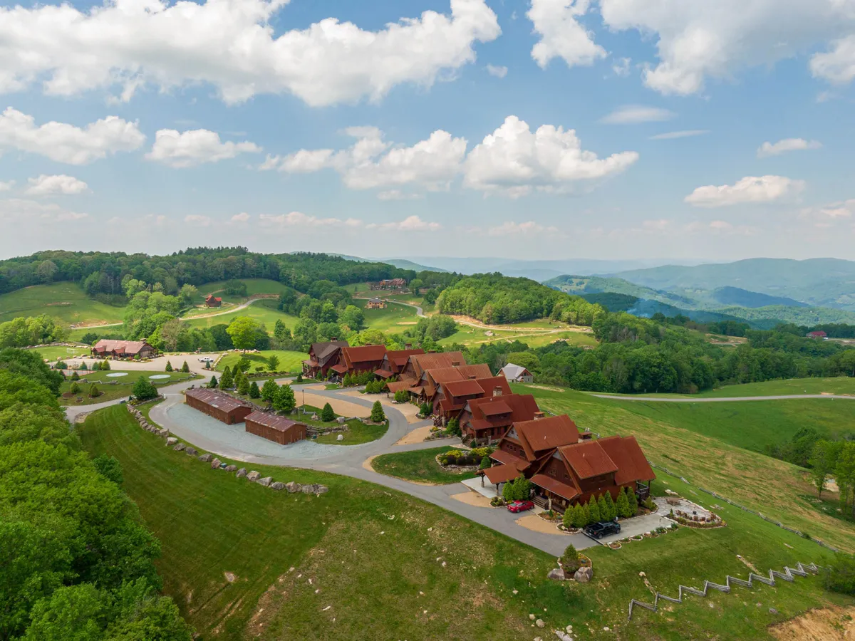 A row of rustic lodges lines a curved driveway, surrounded by lush, green hills and set under a clear blue sky. The accommodations blend seamlessly with the scenic landscape, hinting at the variety of resort amenities available and easy access to the outdoors.