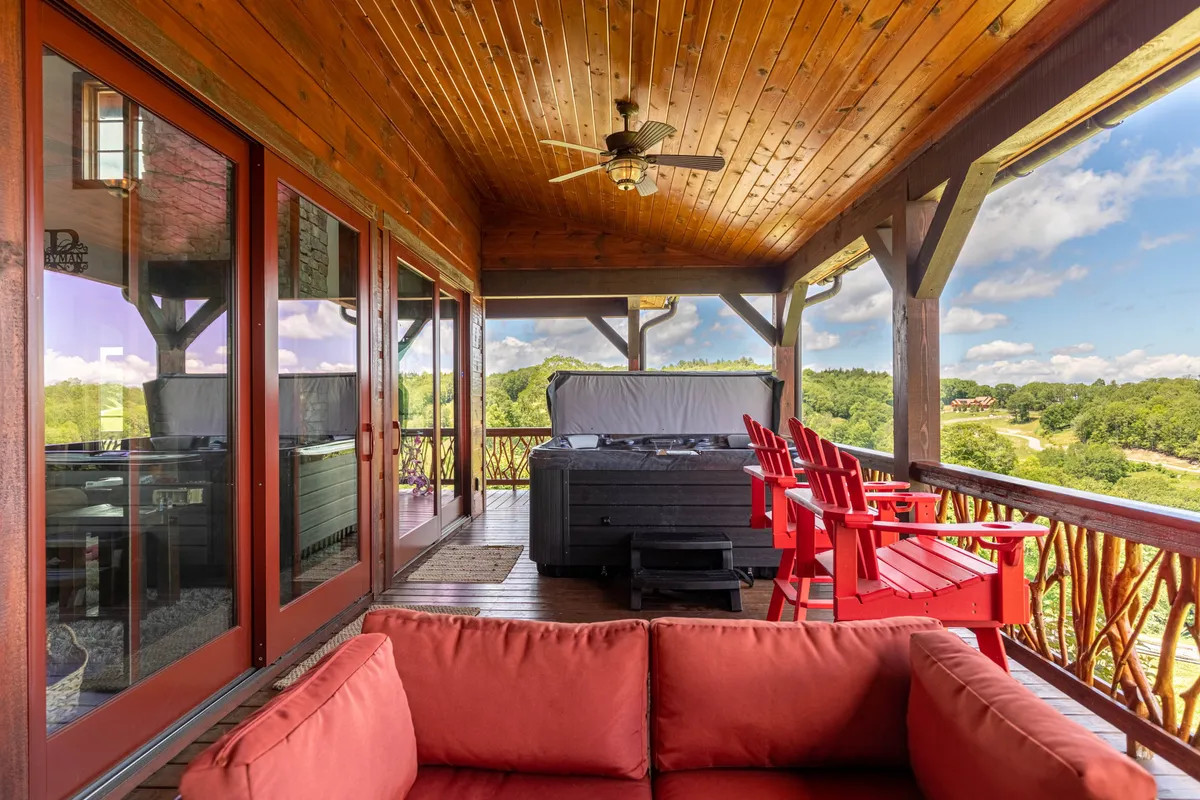 This shot captures a covered deck area with a hot tub, red Adirondack chairs, a wooden ceiling, and large windows providing views of the outdoors. The setting communicates comfort and sociability, inviting guests to relax in the hot tub or gather for conversation, all while enjoying the fresh air and scenic surroundings.