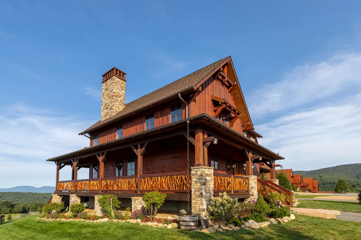 The image showcases a charming two-story wooden cabin with a spacious balcony and a striking stone chimney, set against a dramatic mountain backdrop. The foreground features a landscaped garden, while the covered deck invites guests to relax and take in the sweeping, uninterrupted views. The scene feels warm and welcoming, suggesting mornings with coffee on the porch and evenings spent stargazing by the outdoor fireplace.