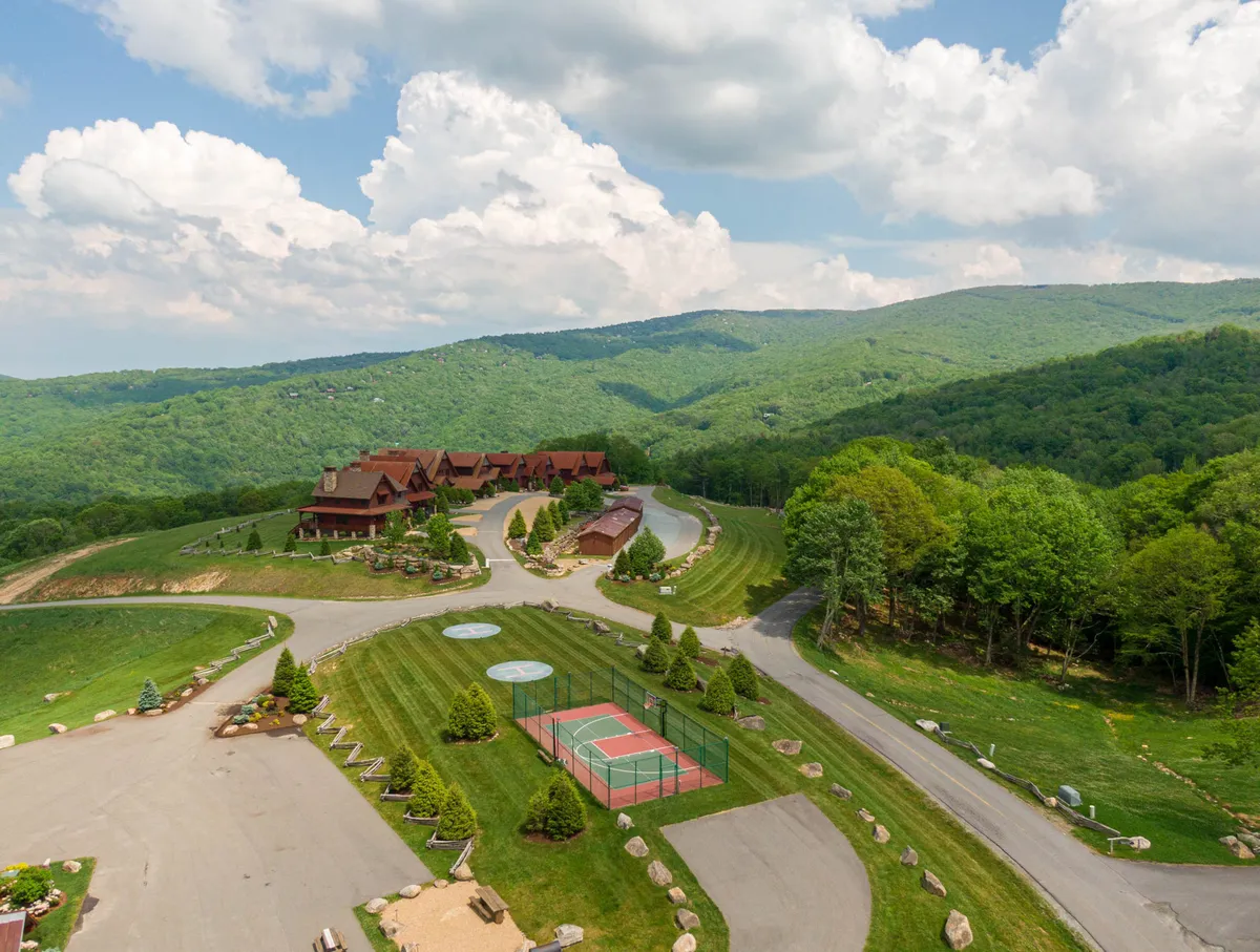 An exterior view of the resort community, highlighting well-maintained grounds, a tennis court, and rustic mountain cabins. The landscape is vibrant and inviting, with clear sightlines to areas for sports, recreation, and relaxation. The setting conveys a sense of active living within a tranquil mountain environment.
