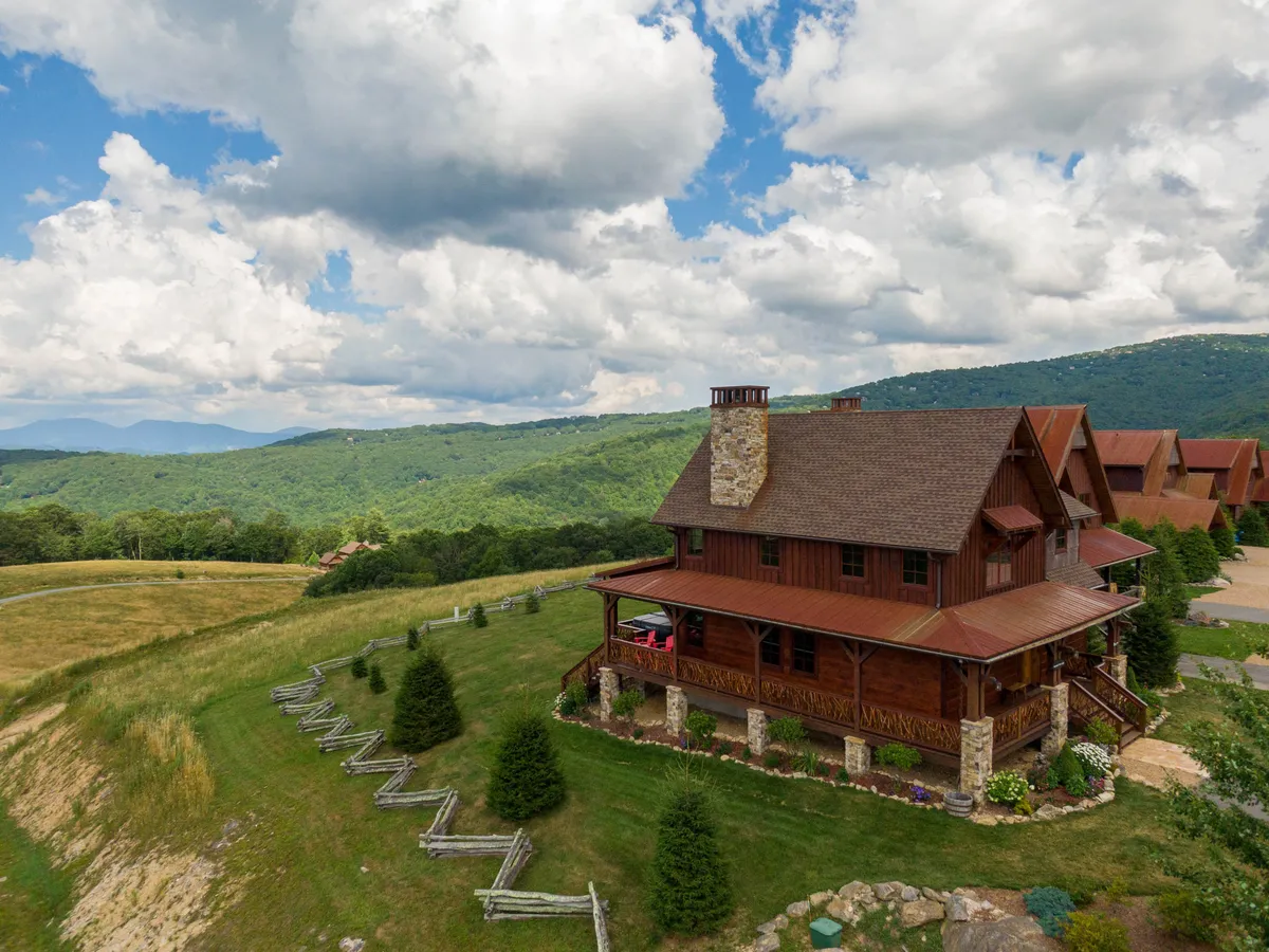 A wide view of a large wooden house with a stone chimney, surrounded by lush green lawn, fenced areas, and dramatic mountain scenery in the background. The home exudes a cozy, welcoming feel, ideal for multi-generational gatherings or groups of friends.