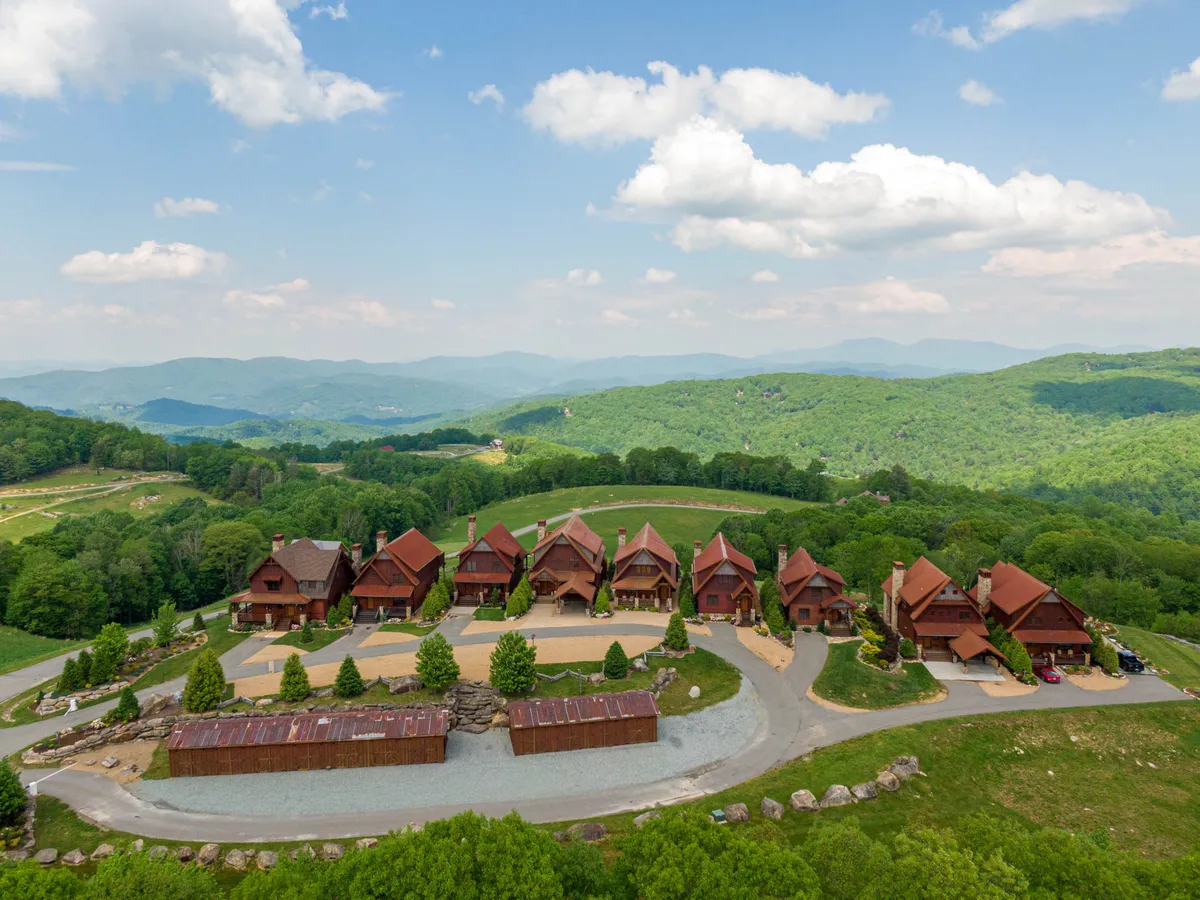 A row of rustic wooden lodges set amidst rich greenery and mountain views under an open sky. The scene suggests a community feel, with multiple accommodations and plenty of space for outdoor activities.